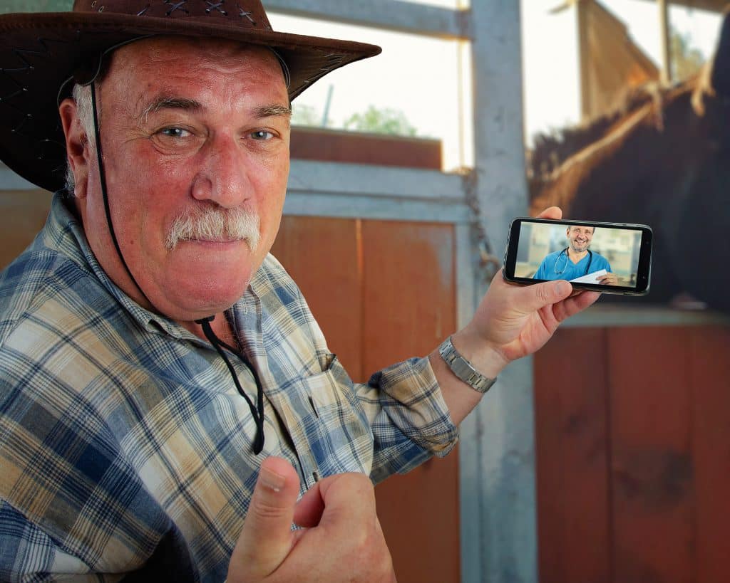 MediStays Care Navigators work closely with regional patients to support their upcoming visit to hospital. Image is of a man wearing a brown oilskin hat and a check shirt holding up a smartphone. He is video calling his doctor while standing next to a wooden stall with a chestnut horse in it.
