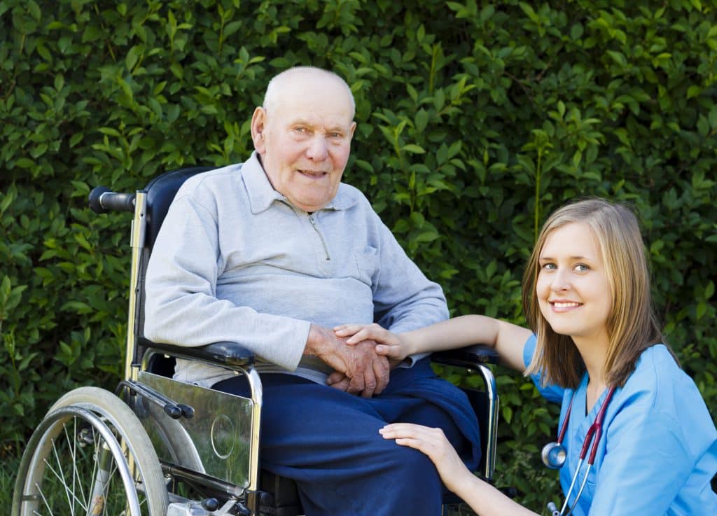 A photograph image of an older man in a manaul wheelchair (left) and a doctor (right). The man is smiling at the camera, with his hands in his lap. He is wearing blue pants and a grey shirt. The doctor to his left has blonde hair to her shoulders and a red stethescope around her neck. She is smiling at the camera and kneeling down next to him. She has her hand place on top of his to support and reassure him. The photograph has been taken outside, in front of a green hedge.