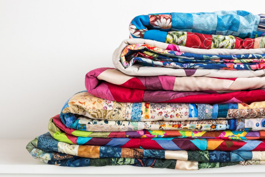 sewing, patchwork and fashion concept - beautiful colorful quilts were neatly folded and stored in several rows in height for storage, sale of finished textile stitched products on white background