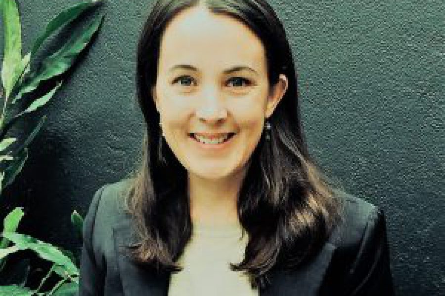 A professional headshot photograph of MediStays Managing Director and Founder Sarah Everitt. Sarah stands against a charcoal wall with a plant in the background. She is wearing a white blouse with a black blazer. She is smiling with long, dark hair around her face.